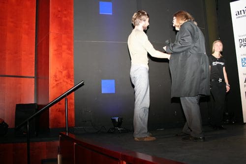 A man in a light sweater shakes hands with a woman in a long black coat on stage. A spotlight is focused on them, with a red and black background visible. Another person stands in the background, observing the interaction.