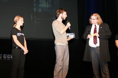 A man receiving an award on stage while holding a trophy, with a woman in a black shirt standing to the left and another man in formal attire presenting the award, all set against a dark backdrop.