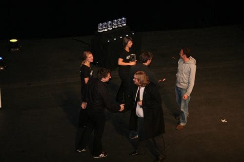 A group of individuals interacting on stage during an event, with some shaking hands. In the background, a table displays multiple trophies, and the stage is dimly lit.