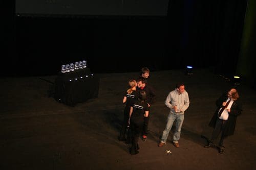 A group of people on stage at an awards ceremony, with two individuals exchanging an award. In the background, several glass trophies are displayed on a black table. An audience member watches from the side.