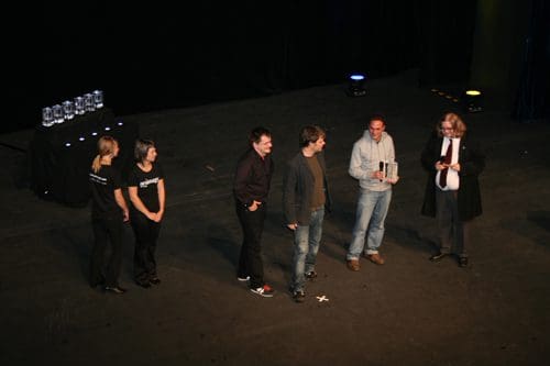 A group of five people stands on a stage, some wearing black t-shirts. They are interacting with each other and holding an award. In the background, several crystal awards are displayed on a table under soft stage lighting.