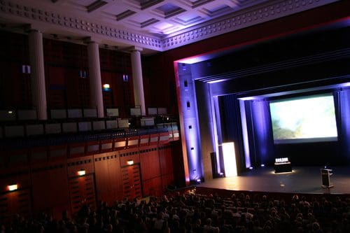 An auditorium filled with an audience facing a stage. The stage features a large screen displaying an image, illuminated by blue lights. Tall columns and an elegant interior design enhance the atmosphere.