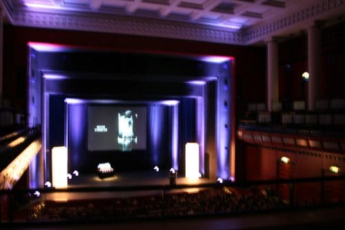A dimly lit theater with a large stage. A projection screen displays monochrome images. The stage is flanked by tall blue and purple lights, creating a dramatic ambiance, while rows of seats can be seen in the foreground.