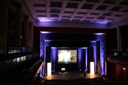 An interior view of a large auditorium featuring a stage illuminated with blue and white lights. The ceiling has a decorative design and columns line the sides. An empty audience area is visible, adding to the spacious atmosphere.