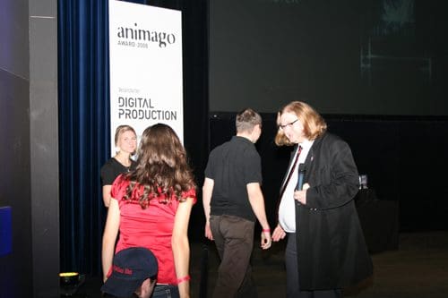 A scene from an award ceremony featuring a banner for "animago Awards 2006". People, including a woman in a red dress and a man in a black coat, are walking across the stage. A spotlight illuminates the area with a dark background.