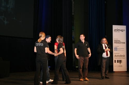 A group of four individuals on stage, engaging in conversation during an event, while a man in a suit observes from the side. The backdrop features a screen and banners of sponsors in a dimly lit auditorium.