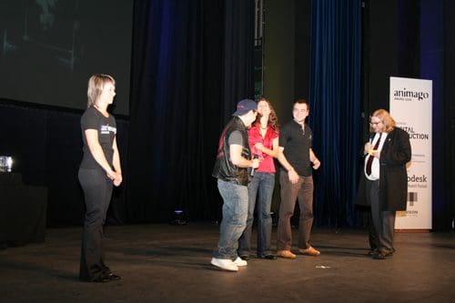 A group of five individuals stand on stage during an event. A woman in a black t-shirt presents from the left, while four others, dressed casually, interact in the center. A large screen and banners are visible in the background.