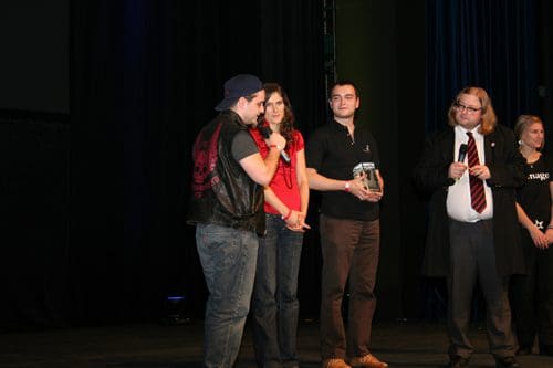 A group of four people standing on stage during an event. One person is speaking into a microphone, while another holds a trophy. A woman in a red shirt looks on, and a man in a suit stands to the side, holding papers.