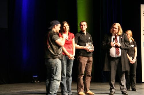 A group of four individuals stands on stage during an award ceremony, with two men speaking and holding a trophy, while the others listen attentively. The backdrop features soft lighting, and audience members can be seen in the darkened area behind.
