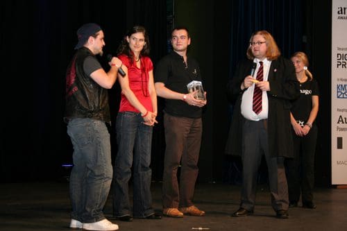 A group of four people on stage during an award ceremony. One person is speaking into a microphone, while another holds a trophy. The audience is slightly blurred in the background, with a backdrop displaying award logos.