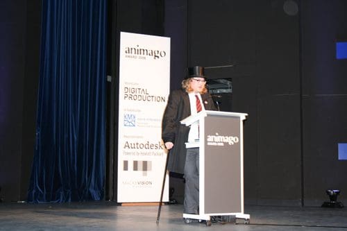 A man in a black suit and top hat stands at a podium delivering a speech at the animago Awards 2008 event. Behind him is a blue curtain and a large sign for animago, with logos of sponsors including Autodesk.