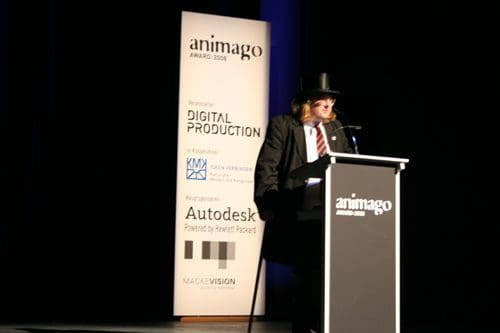 A presenter wearing a black suit, white shirt, and top hat stands behind a podium at the animago awards, with a spotlight highlighting the stage. A banner displays event sponsors and the animago logo in the background.