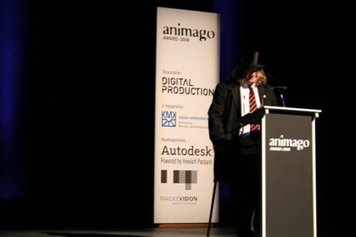 A man wearing a top hat and a blazer stands at a podium during the animago awards ceremony. Behind him, a banner displays the event's sponsors and logo, set against a dark stage backdrop.