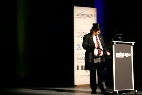 A presenter wearing a suit and a top hat stands beside a podium on a stage, with colorful lighting in the background. The backdrop features signage for an award ceremony, creating a formal atmosphere.