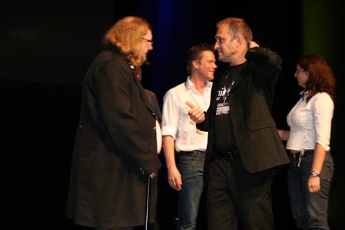 A group of individuals interacting on stage during an event, with one person in a long black coat and another in a black shirt. The backdrop is dark, highlighted by colorful stage lighting.