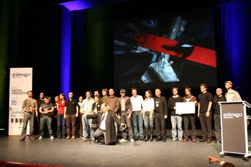 A group of people standing together on a stage during an event, with a large screen in the background displaying an abstract image. The stage is adorned with colorful lighting, and some attendees hold awards while others pose for a photo.