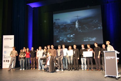 A diverse group of people stands together on stage, posing for a photo during an event. In the background, a large screen displays an aerial scene with clouds and an airplane. The stage is decorated with banners from various sponsors.