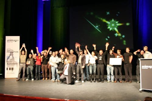 A large group of professionals celebrating on stage at an event, with many individuals waving their hands. The background features colorful lighting and a digital display, adding a festive atmosphere to the scene.