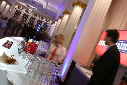 A stylish event space featuring a sleek bar with glasses and drinks. A blurred figure in a white shirt and red tie serves cocktails. Elegant columns and dim lighting create a sophisticated atmosphere.