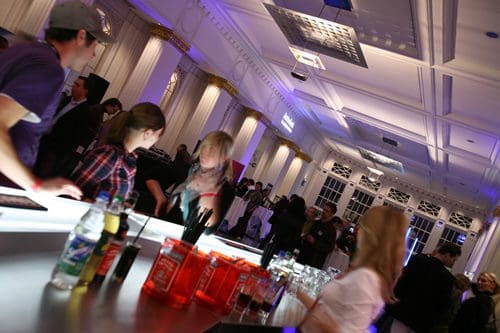 A lively cocktail bar scene in a spacious venue, featuring smiling patrons interacting while standing at the bar. The bar is lined with various colorful bottles, and the elegantly lit room has decorative pillars and a festive atmosphere.