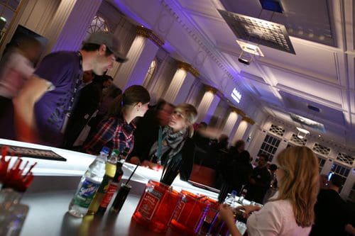 A busy bar scene featuring patrons interacting and ordering drinks. Two men converse near the bar, while a woman with a scarf chats with a bartender. Colorful bottles line the countertop, with soft lighting creating an inviting atmosphere.
