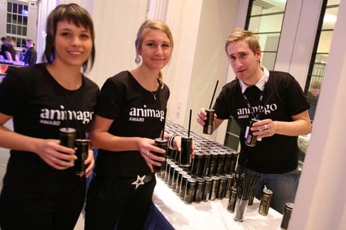 Three hosts wearing black shirts with "animago AWARD" logo, smiling while holding cans. They are in front of a table stacked with energy drink cans, set in a lively event atmosphere.