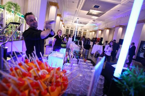 A bartender shaking a cocktail behind a bright bar scattered with colorful fruit garnishes. In the background, guests socialize in an elegantly decorated event space with soft lighting and white tablecloths.
