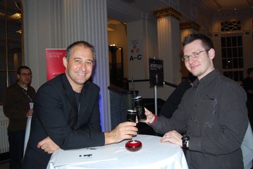 Two men smiling and clinking glasses at a table during an event. The backdrop features elegant pillars and LED lighting, creating a festive atmosphere. One man wears a black jacket while the other is in a black shirt.
