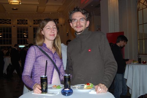 A woman in a purple sweater and a man in a gray sweater standing together at a social event, each holding drinks. They smile at the camera, with a table featuring plates of food in the foreground and elegantly decorated surroundings in the background.