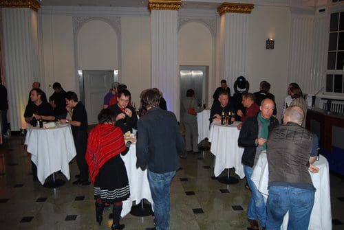 A gathering of people socializing around high tables in a well-lit event space with elegant columns. Guests are enjoying drinks and snacks, wearing a mix of formal and casual attire, creating a lively atmosphere.