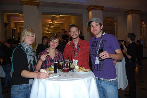 Four young adults gathered around a high table at an event, enjoying food and drinks. They are casually dressed, with a woman in a scarf and a man in a purple shirt smiling for the camera. The background features an elegant venue.