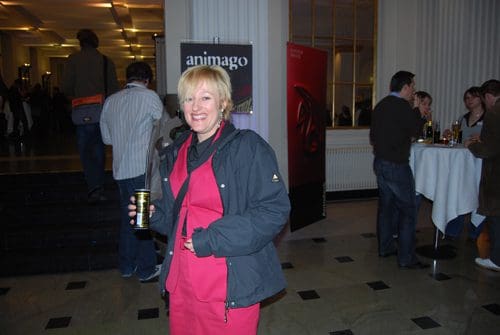 A smiling woman in a pink outfit holding a drink, standing in a crowded event space. In the background, people are gathered around tables, and banners display event branding.