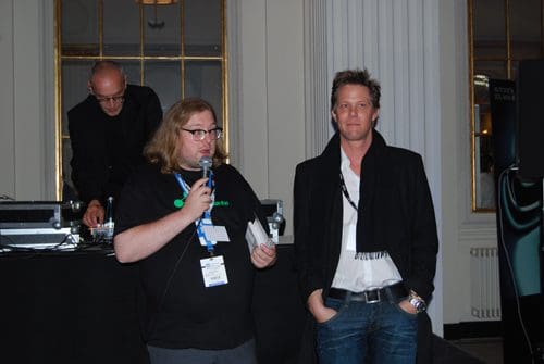 A man with long hair holding a microphone speaks to another man wearing a black scarf at an event. In the background, a DJ operates equipment. The setting is elegantly decorated with large windows.