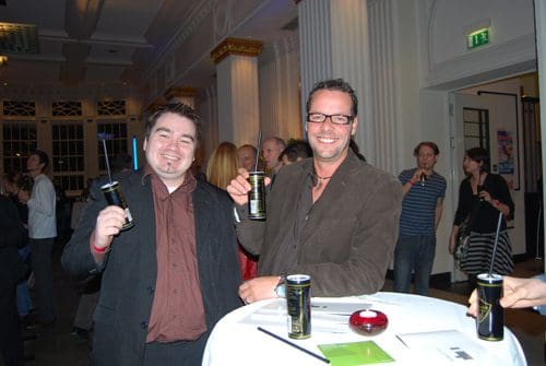 Two smiling men hold cans of energy drink while standing at a round table in an elegant event space. The background features guests mingling and a bright, festive atmosphere.