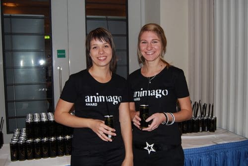 Two women wearing black t-shirts with the "animago Award" logo, smiling while holding drinks. Behind them, cans are neatly arranged on a table in a well-lit event space.