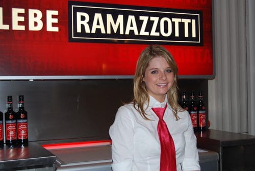A woman with long blonde hair stands smiling in front of a bar display. She wears a white shirt and a red tie, with bottles of Ramazzotti on the counter behind her. A bright red sign featuring the brand name is visible in the background.