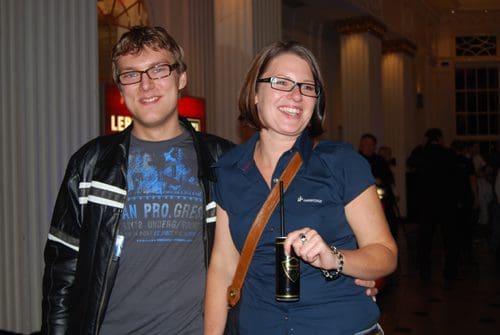 A smiling man and woman stand together in a spacious venue with elegant columns. The man wears glasses and a black jacket over a blue shirt, while the woman holds a can in one hand and wears a blue shirt with a brown strap bag.