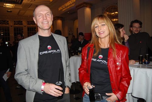 Two individuals at a social event, wearing matching black t-shirts featuring the word 'CREAPOLIS.' One person, a man, is smiling and wearing a light gray blazer, while the woman beside him has long blonde hair and is dressed in a red leather jacket. They are both holding drinks, and the backdrop features blurred figures and decorations.