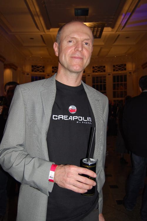 A man wearing a gray blazer over a black shirt with 'CREAPOLIS' printed on it, holding a can with a straw. He is posing in a well-lit indoor venue filled with people in the background.