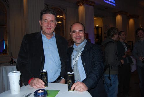 Two men standing together at a social event, smiling while holding cans of energy drink. The background features a crowd of attendees and elegant decorations in a well-lit venue.