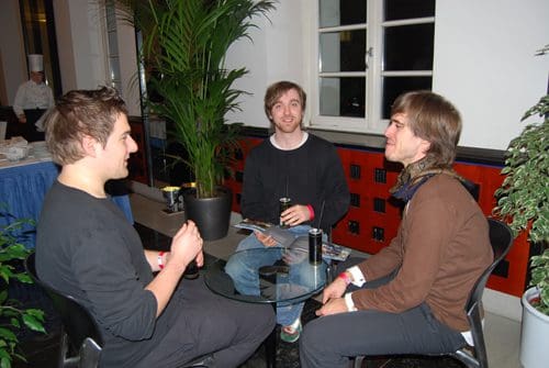 Three young men sitting around a small glass table in a well-lit indoor space. They are engaged in conversation, with one man holding a drink. A large potted plant is placed nearby, and there is a buffet setup in the background.