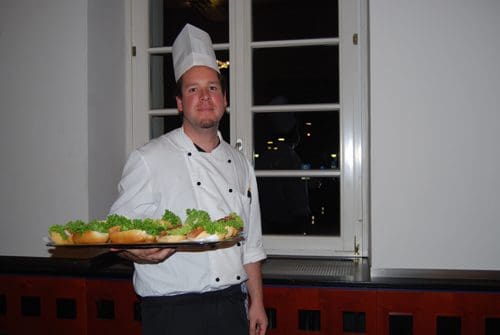 A chef wearing a white uniform and hat stands holding a tray of sandwiches topped with fresh lettuce, in a well-lit dining area near a window.