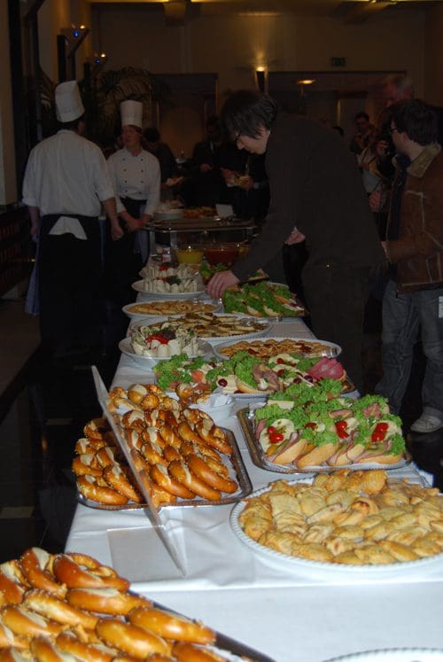 A buffet table filled with an array of dishes including sandwiches, pastries, and salads. People are gathered around, with a chef in the background, as guests serve themselves from the spread.