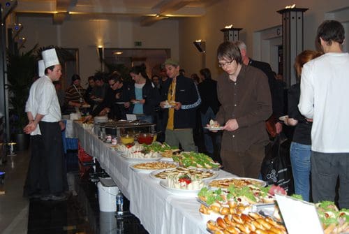 A buffet spread featuring various dishes on a long table with guests serving themselves. A chef in a white hat stands at one end, while several people enjoy the food in a well-lit event space.