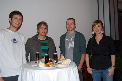 Four young adults standing around a high table with food and drinks. They are dressed casually, smiling, with plates of appetizers and beverages on the table, set against a plain backdrop.