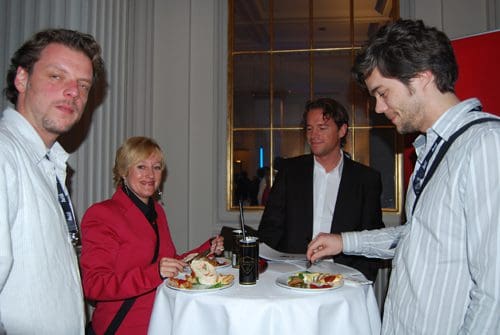 A group of four people standing around a high table in a well-lit room. Two men and a woman are eating light meals from plates, while another man observes. The woman wears a red blazer, and the atmosphere is casual.