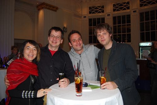 A group of four friends posing together at a social event, standing around a round table. They all hold drinks, smiling. The venue features high ceilings and elegant decor, creating a lively atmosphere.