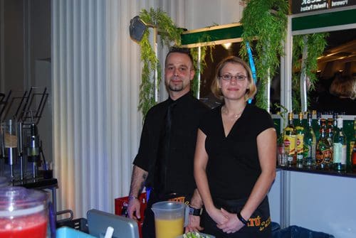 A bartender and a bar manager pose behind a well-stocked bar. The male bartender wears a black shirt and tie, while the female manager is in a black blouse. The bar displays various bottles of liquor and fresh greens in the background.