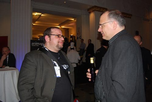Two men engaged in a conversation at an event, with one holding a bottle. A dimly lit ambiance highlights a gathering atmosphere, featuring tables and other attendees in the background.