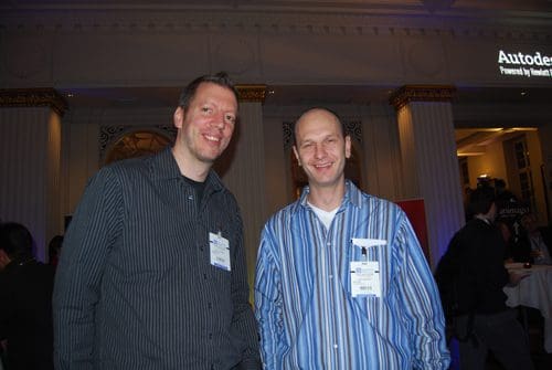 Two men smiling and standing together at a social event. One wears a black striped shirt, while the other is in a blue and white striped shirt. The background features elegant decor and dim lighting, common in formal gatherings.